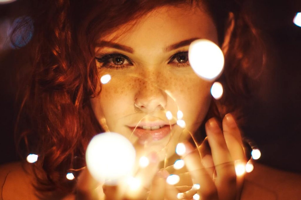 A close-up photo of a vibrant red-headed woman holding a string of white LED lights. 